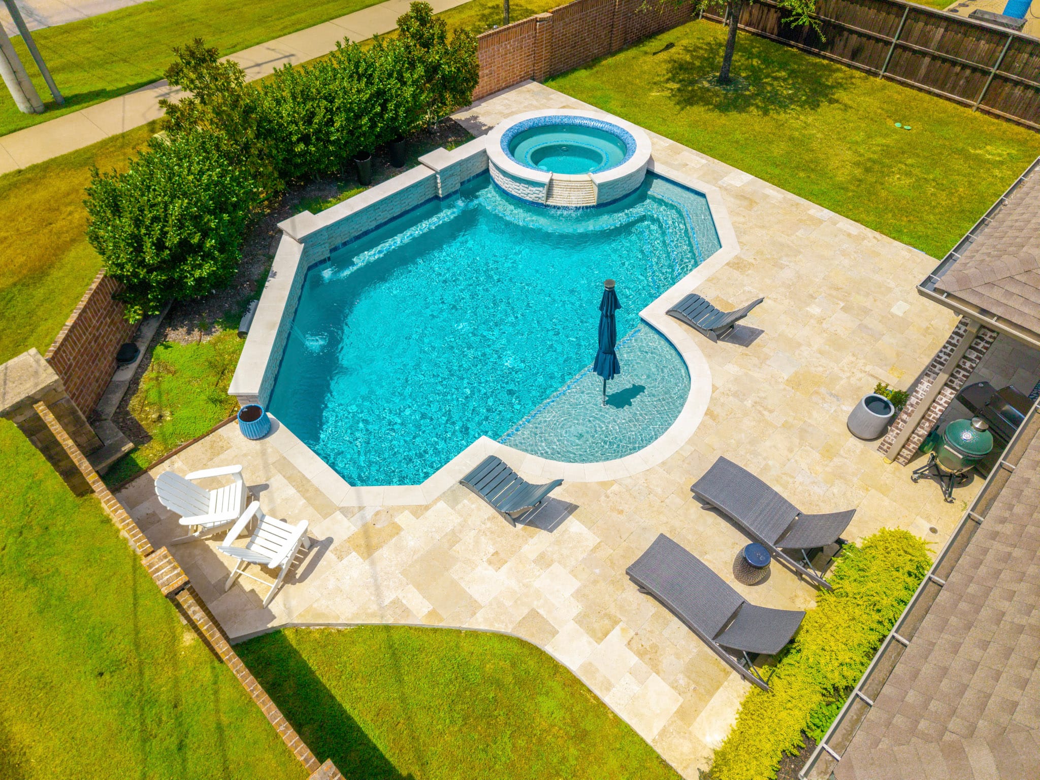 Angled drone view of a Frisco pool remodel with raised spa, sun shelf, water features and travertine decking.