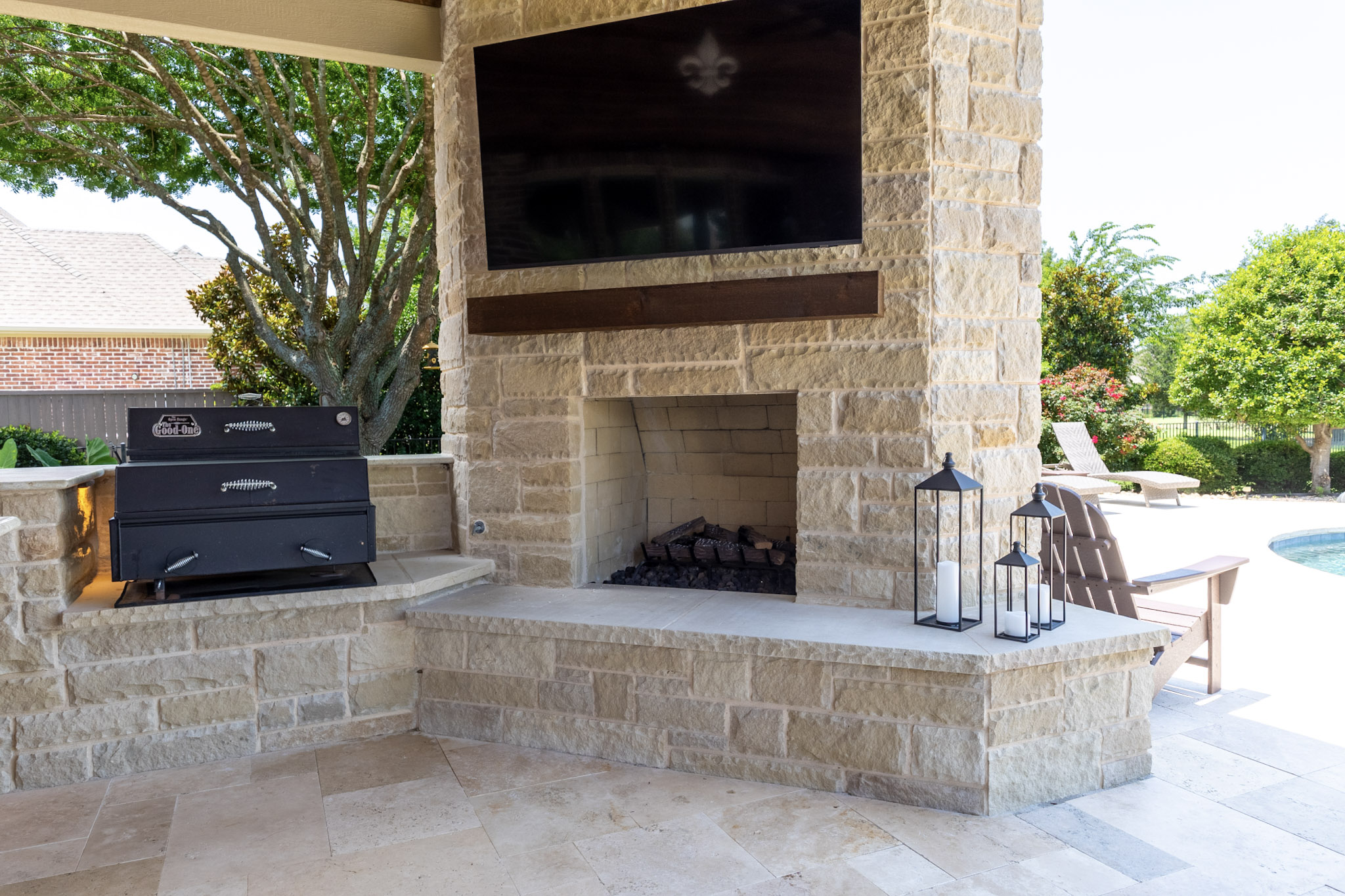 Stone outdoor fireplace with mounted television, wood mantel, hearth, and adjacent cooking equipment.