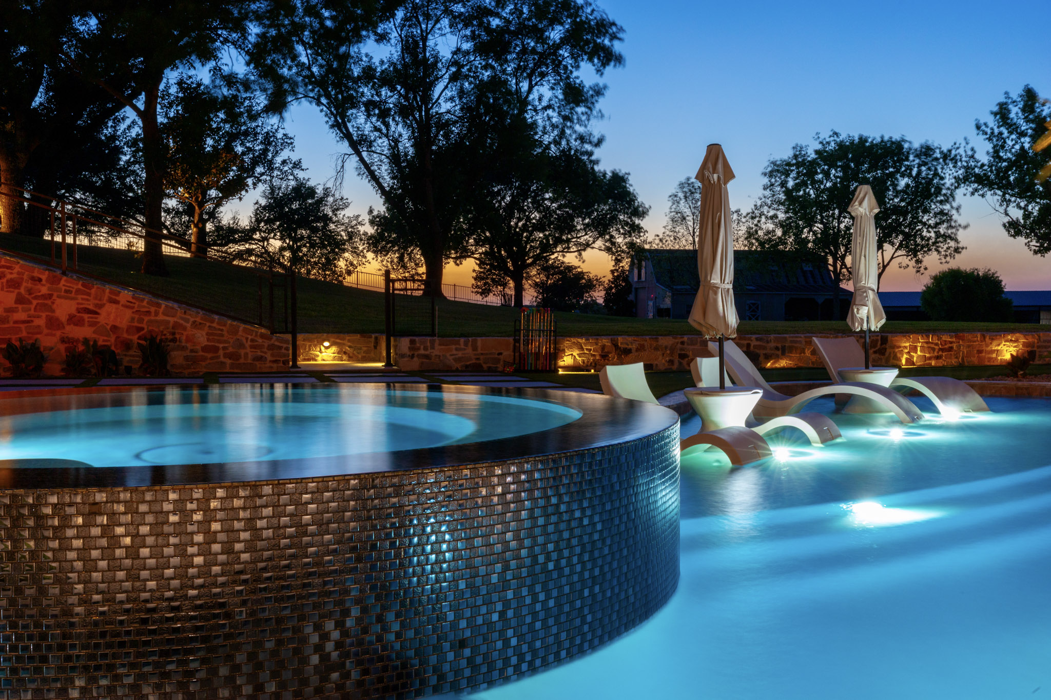 Glass-tile round spa beside illuminated submerged pool loungers.