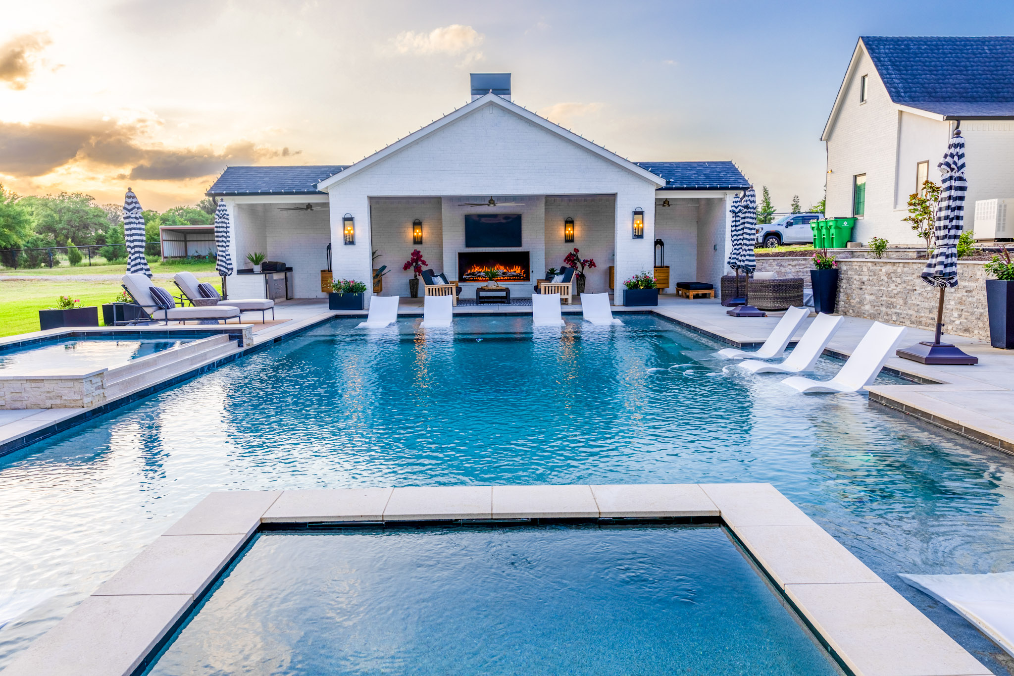 Large geometric swimming pool leading toward a covered fireplace pavilion at sunset.