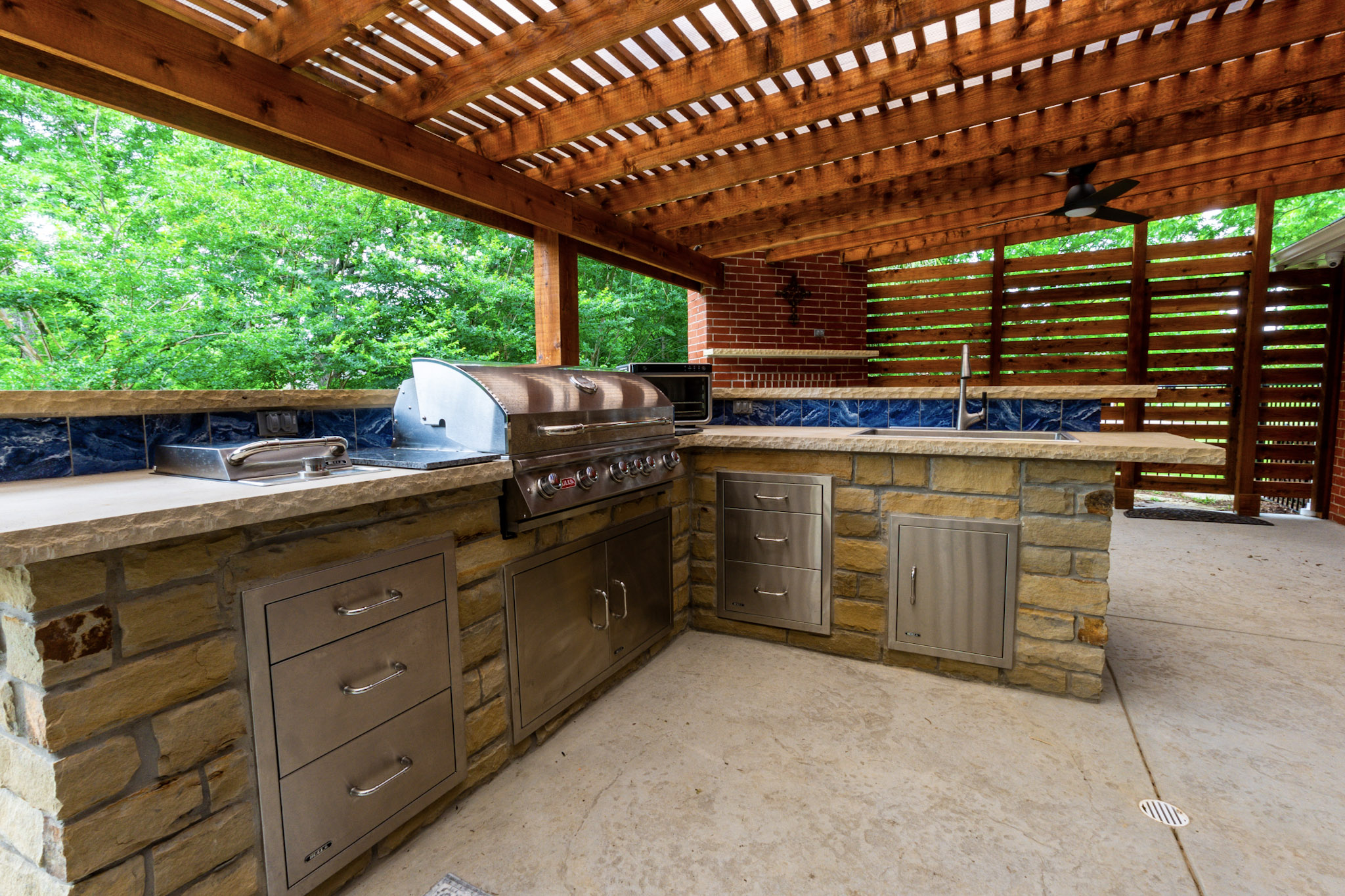 Stone outdoor kitchen with stainless grill, sink and storage beneath a wood pergola.