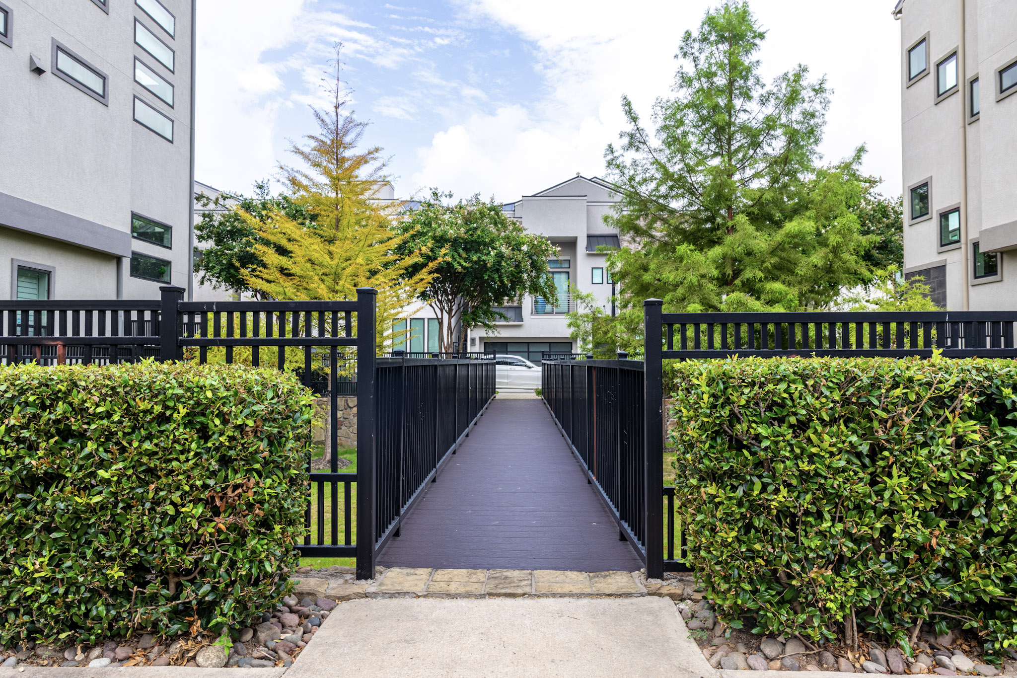 Pedestrian bridge with black railings surrounded by hedges and trees.