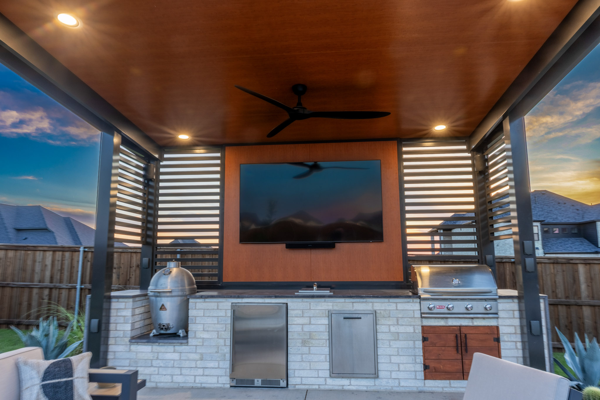 Covered outdoor kitchen with built-in grills, television and modern wood ceiling.