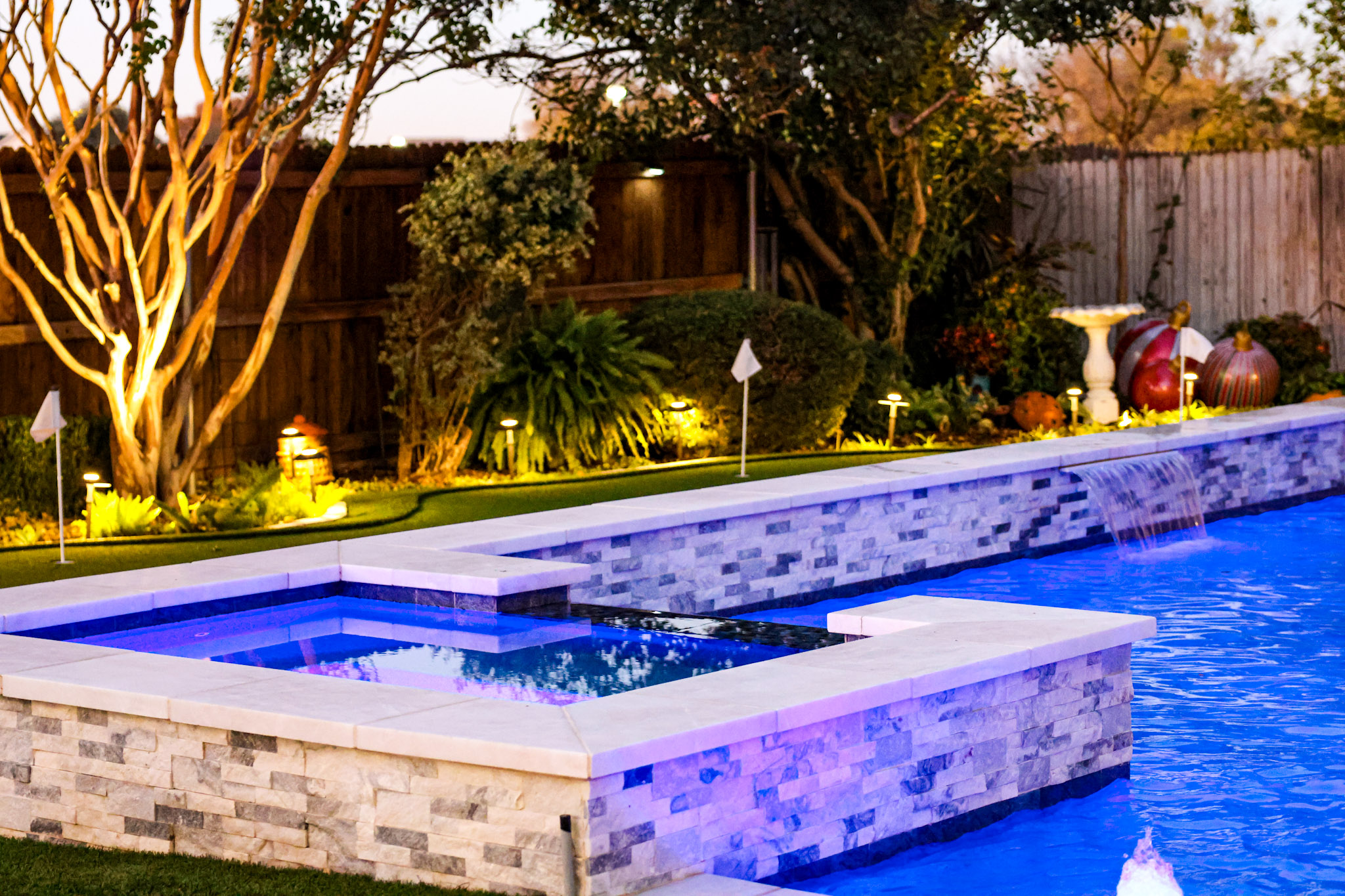 Raised square spa with white coping and stacked-stone exterior beside an illuminated blue pool.