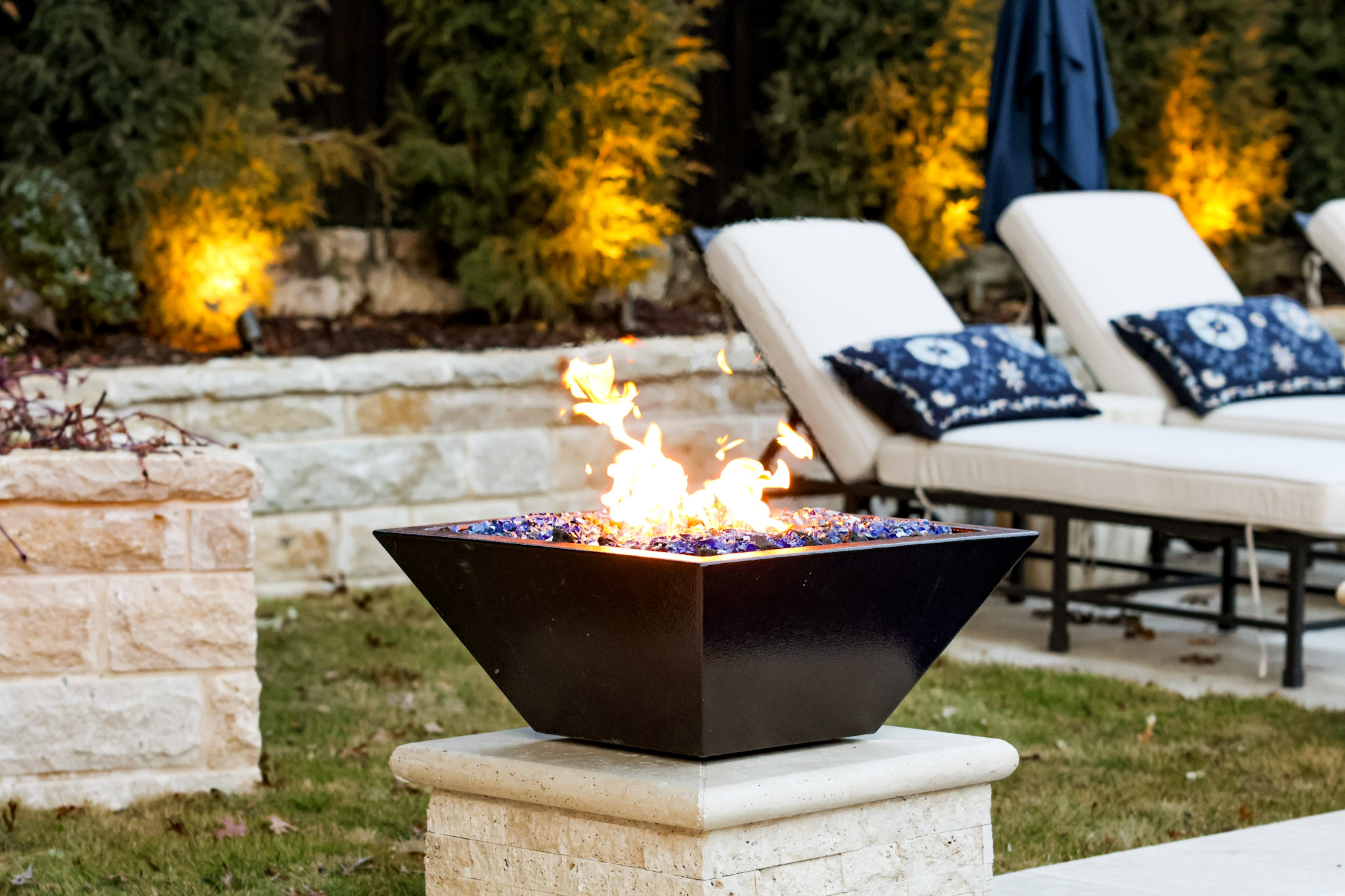 Black fire bowl with flames beside poolside lounge chairs.