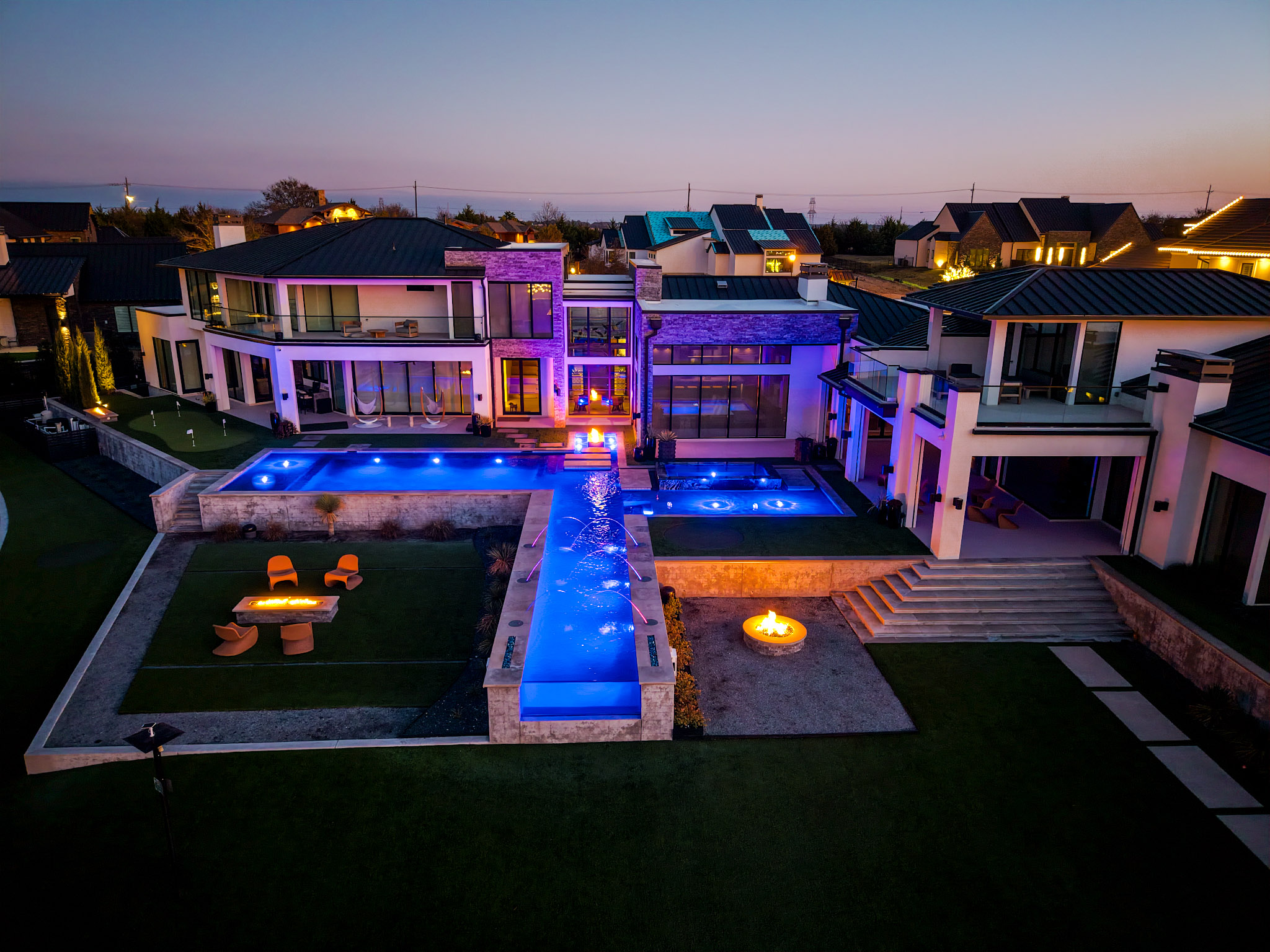 Blue-hour aerial of illuminated luxury pool and outdoor living areas.