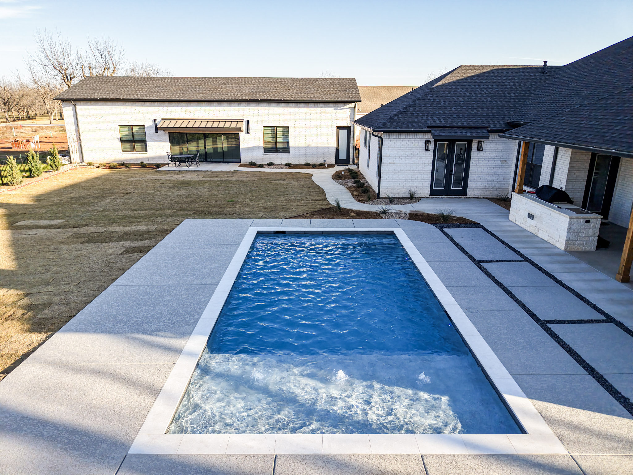 Aerial view of a rectangular pool with tanning ledge, concrete deck and adjacent home.