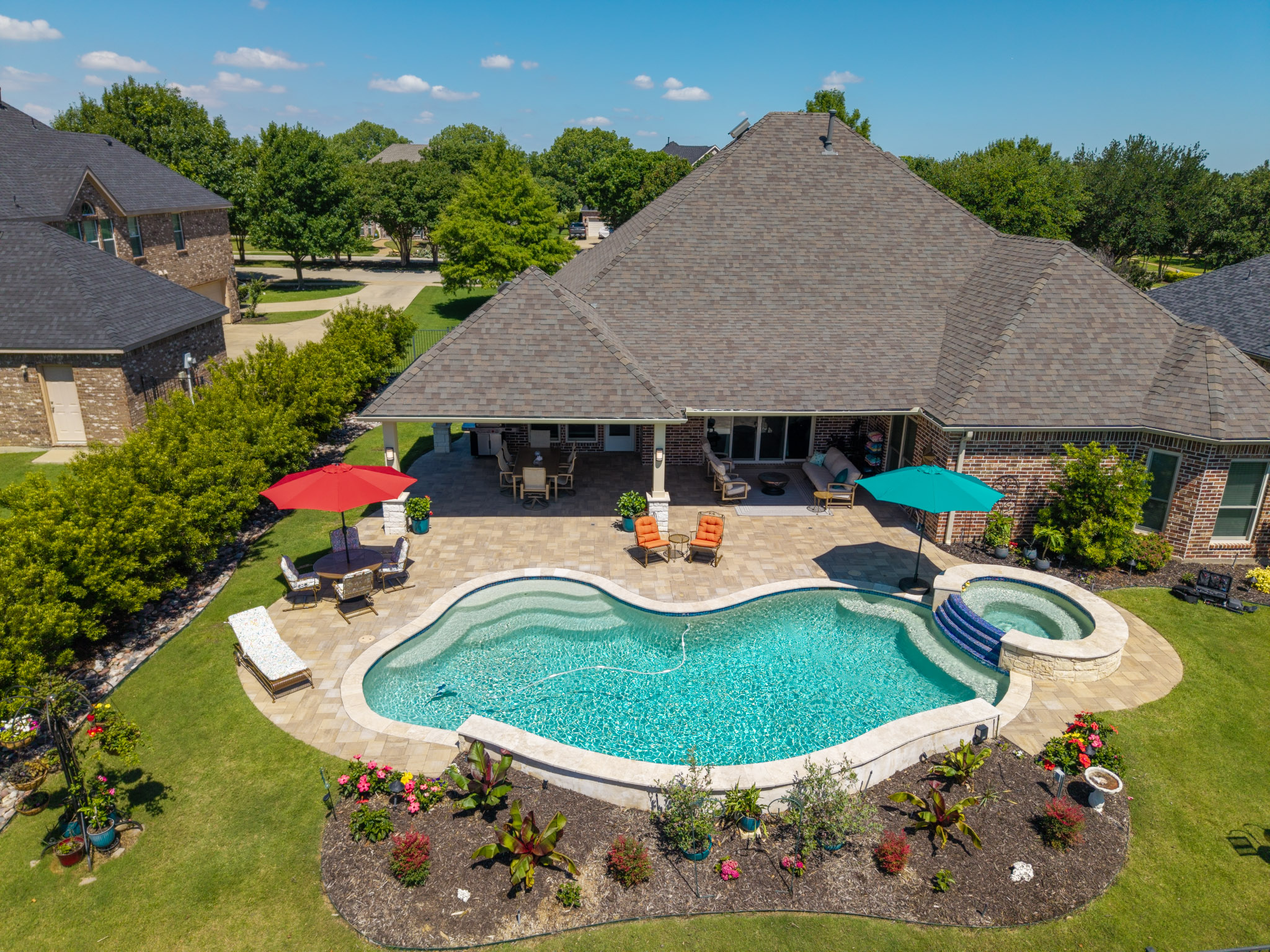 Wide aerial of brick home with extended covered patio, freeform pool, spa, paver deck and landscaping.