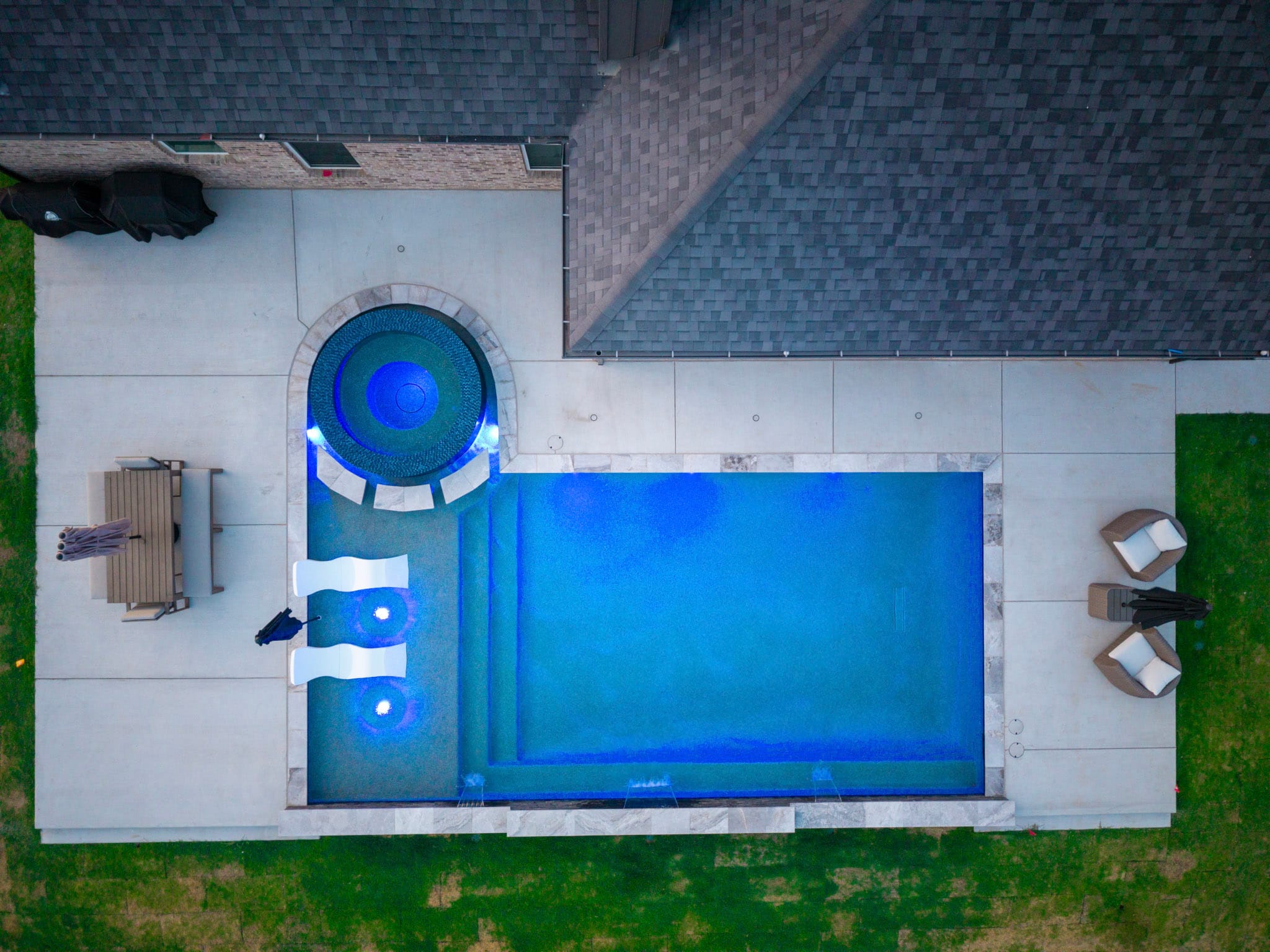Straight-down nighttime drone view of a blue-lit geometric pool, circular spa and tanning ledge.