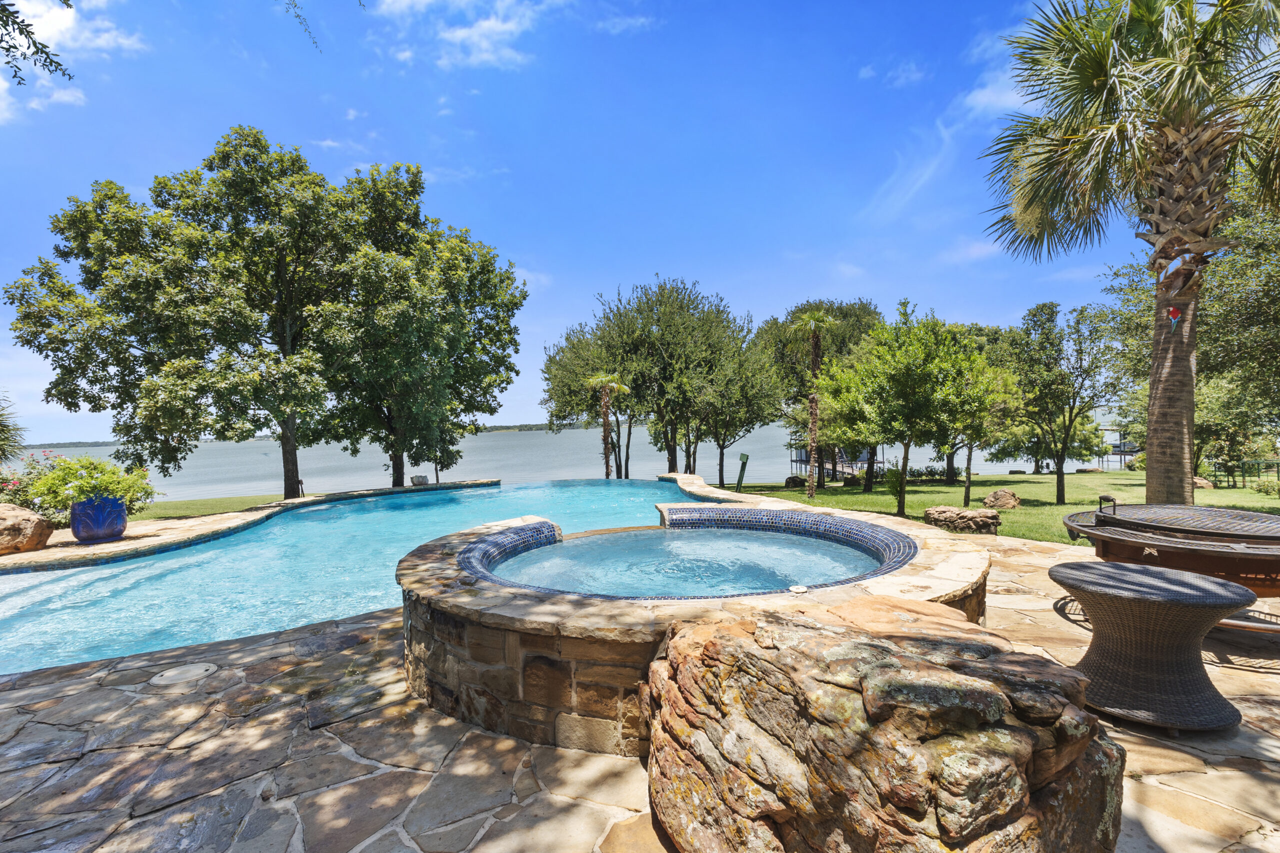 Circular stone spa beside a blue freeform pool with a lake visible beyond.