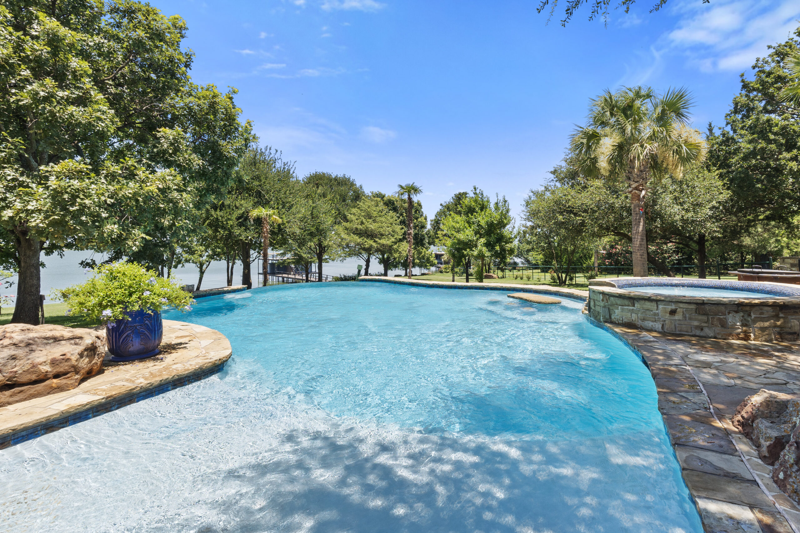 Freeform blue swimming pool surrounded by stone, mature trees and palm trees near a lake.