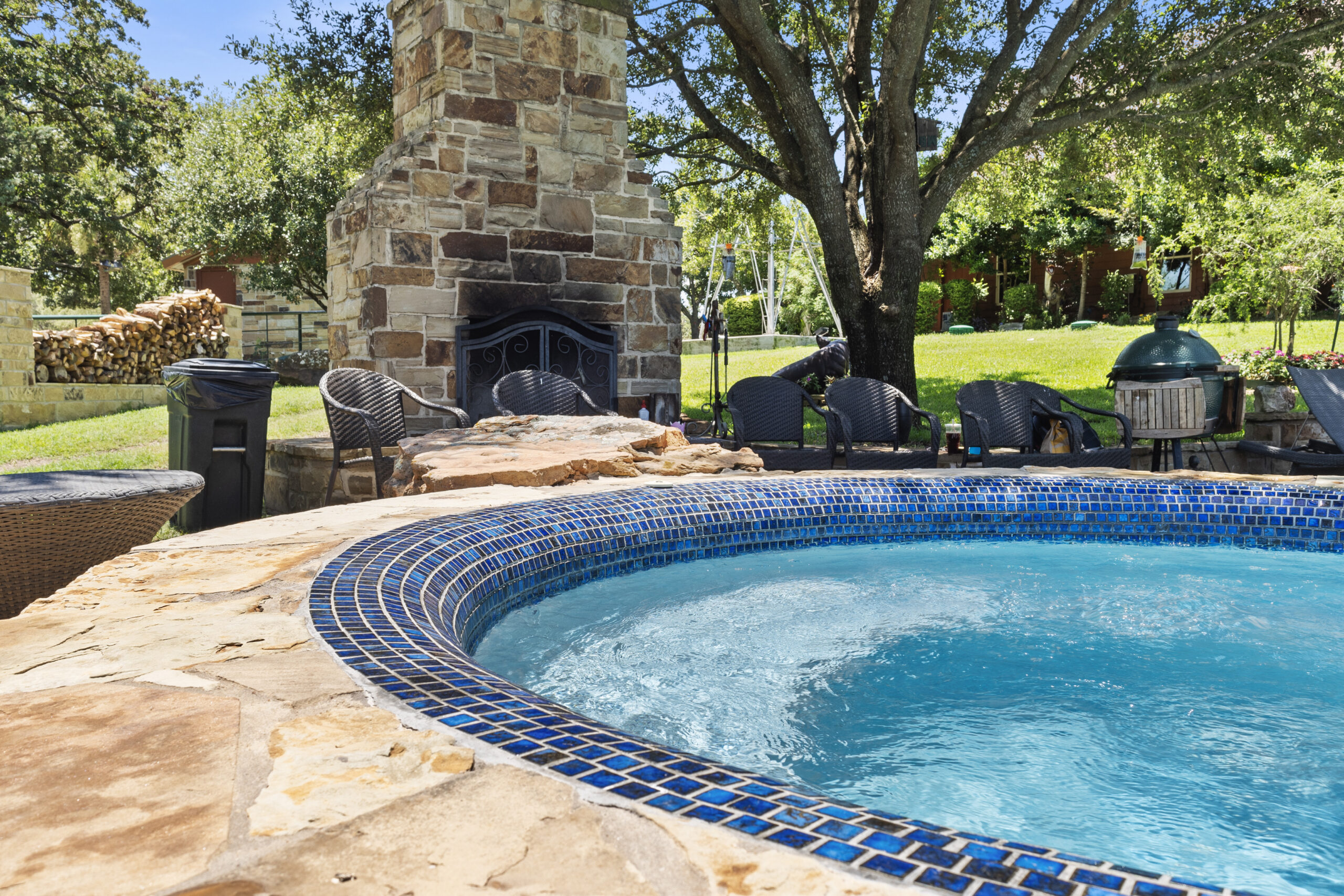 Circular spa with blue mosaic tile and natural-stone coping beside an outdoor fireplace.