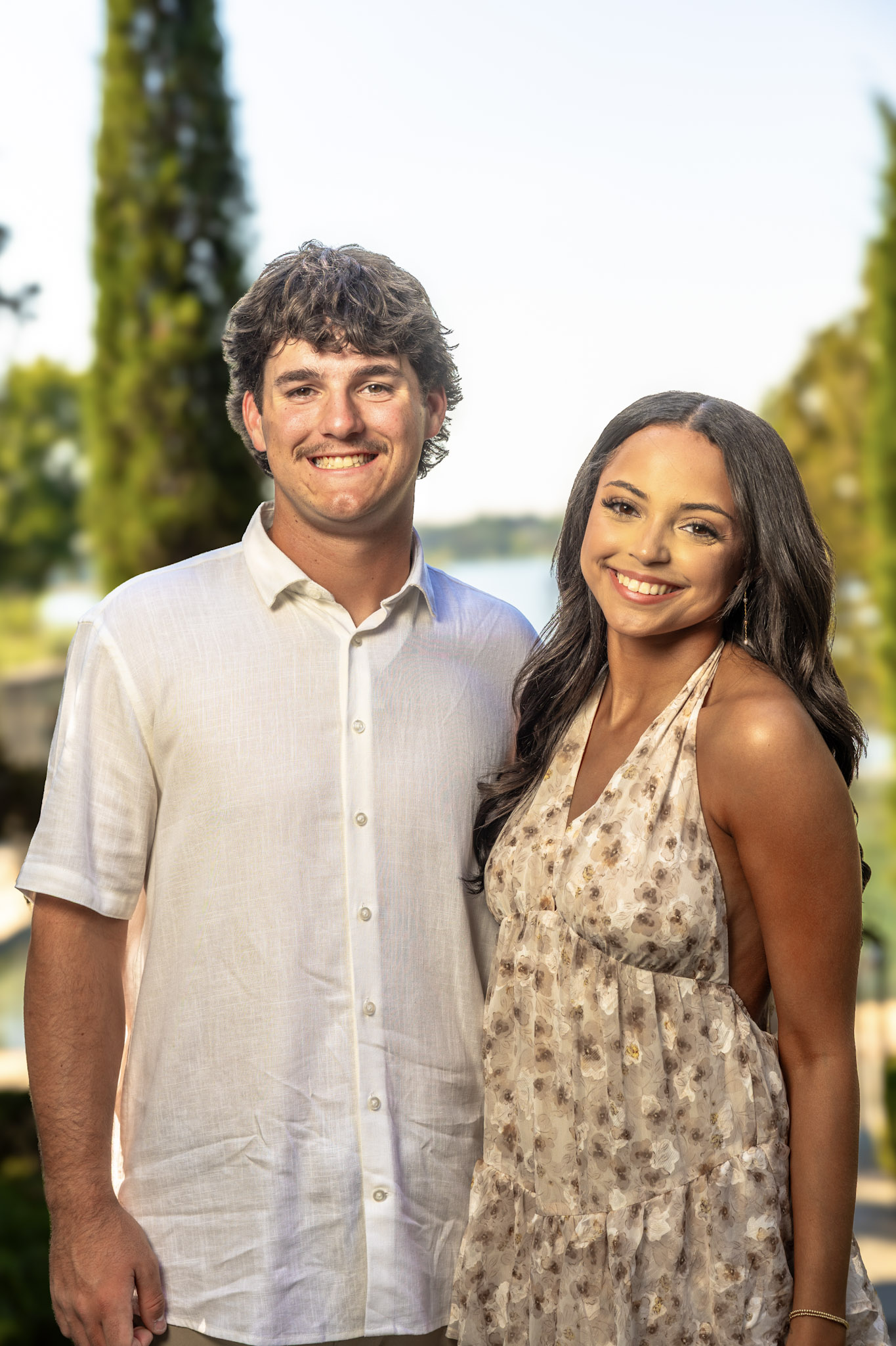 Engaged couple posing together with trees and water softly blurred behind them