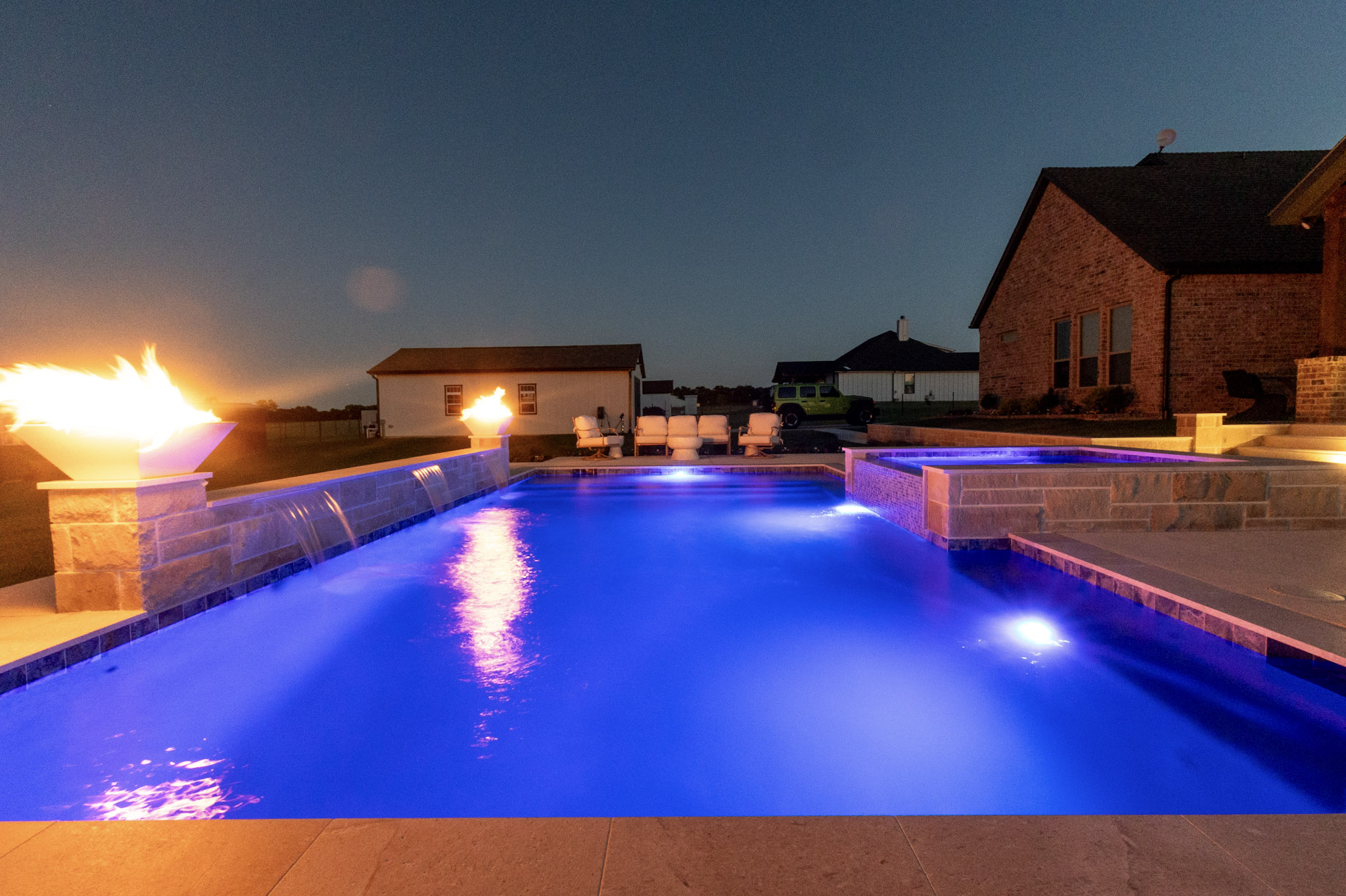 Blue illuminated swimming pool with fire bowls and raised spa at night